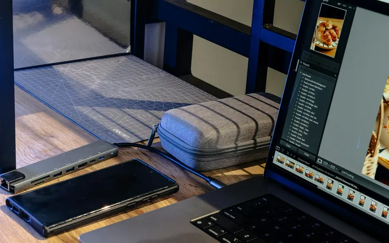 Computador, telefone e acessórios sobre uma mesa de madeira.
